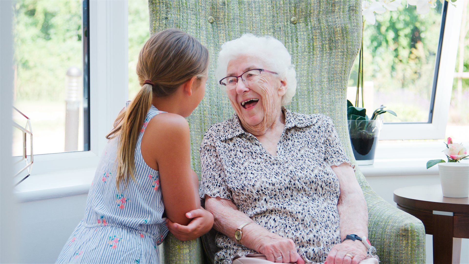 Granddaughter Sitting And Talking With Laughing Grandmother During Visit To Retirement Home