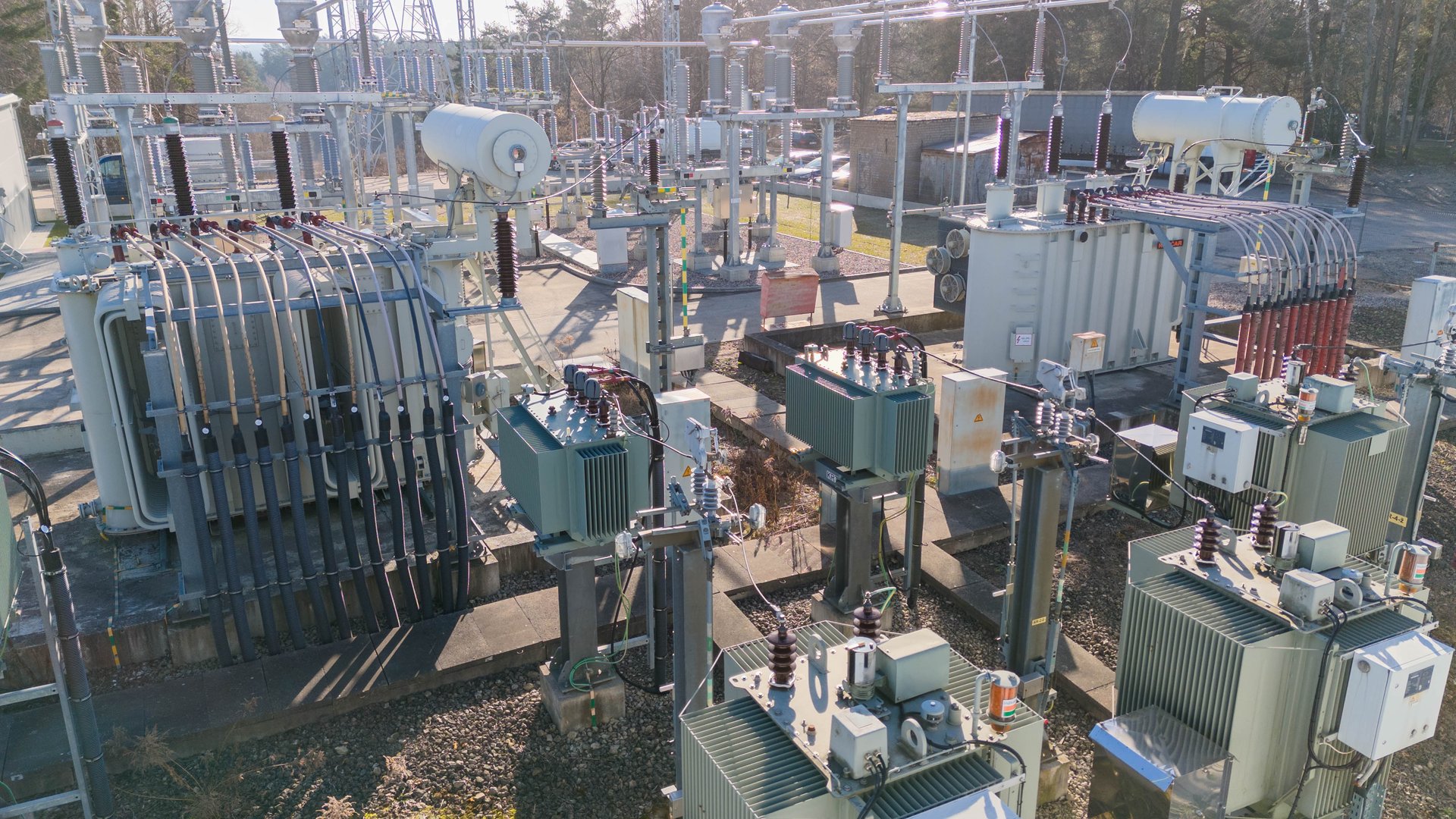 An industrial electrical substation featuring various transformers, circuit breakers, and power lines. The scene is set outdoors with clear skies and sunlight illuminating the equipment.