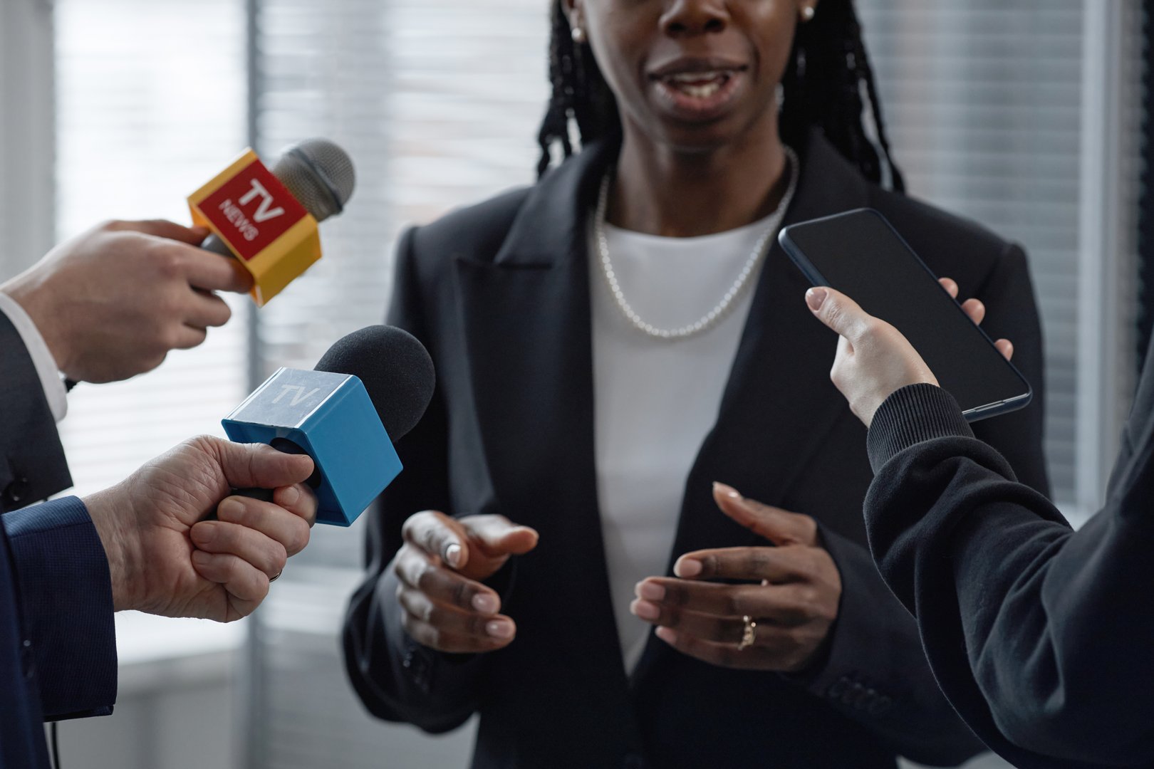Cropped shot of Black female politician gesturing with hands answering questions, while speaking to group of journalists holding microphones and smartphone during press conference in office