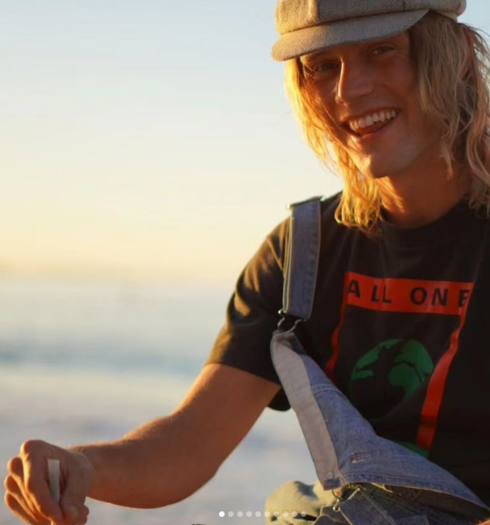Person with long hair and a cap smiling, wearing a black shirt and overalls, sitting outdoors with blurred background.