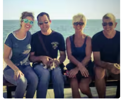 Four people sitting on a bench by the sea, with one holding a small dog. It's a sunny day.