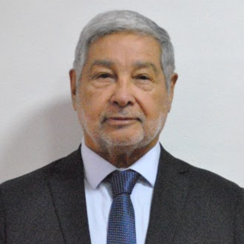 Elderly man with gray hair and beard wearing a dark suit, light shirt, and patterned tie, standing against a plain background.