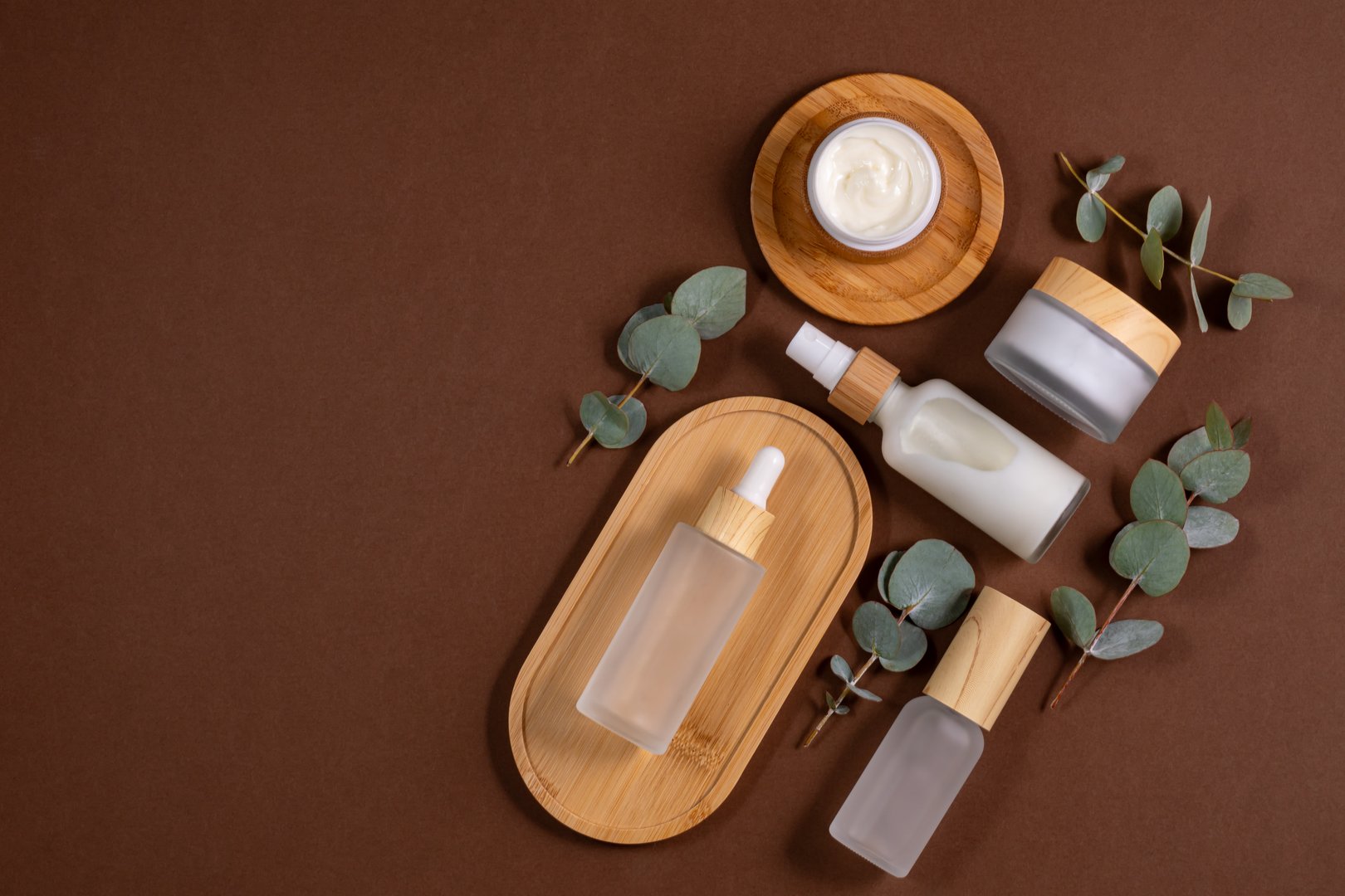 Frosted glass bottles and jars with natural cosmetics and eucalyptus on wooden tray on table in bathroom. SPA organic bio cosmetics set. Space for text.
