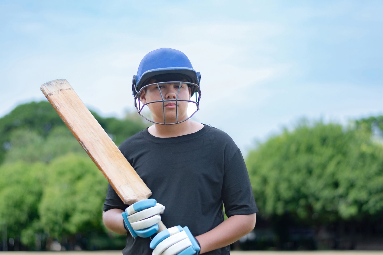 Young cricket sport player wears helmet and gloves, holding cricket bat and old cricket ball in outdoor field, soft focus, hobbies and education concept.