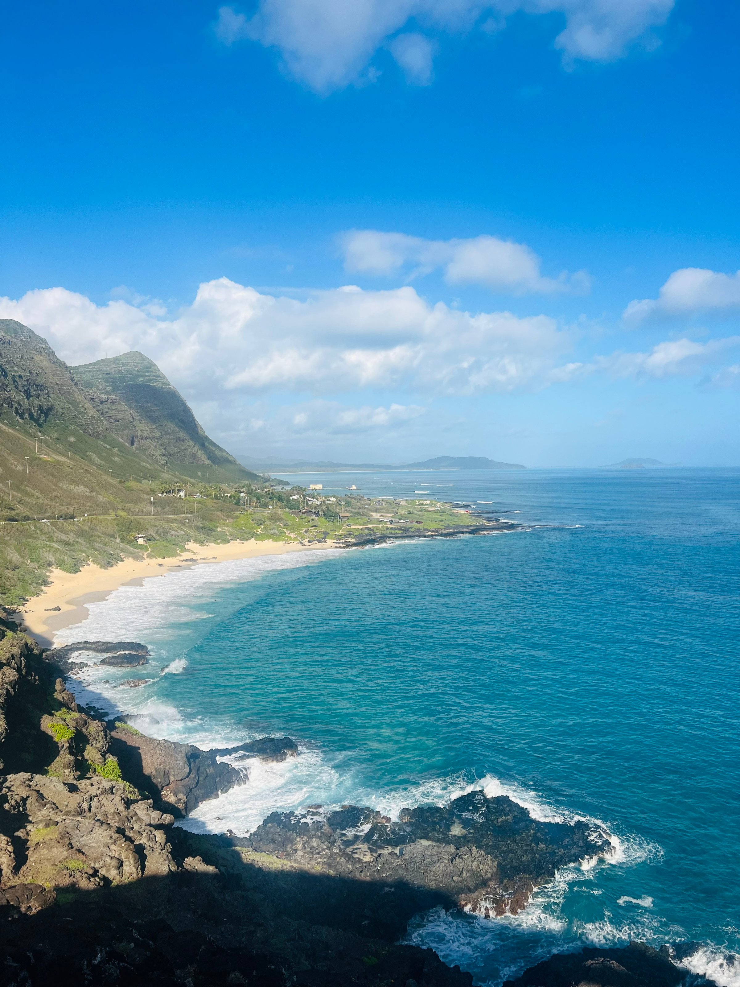 A scenic coastal view with a sandy beach, rocky shoreline, and blue ocean under a clear sky with fluffy clouds.