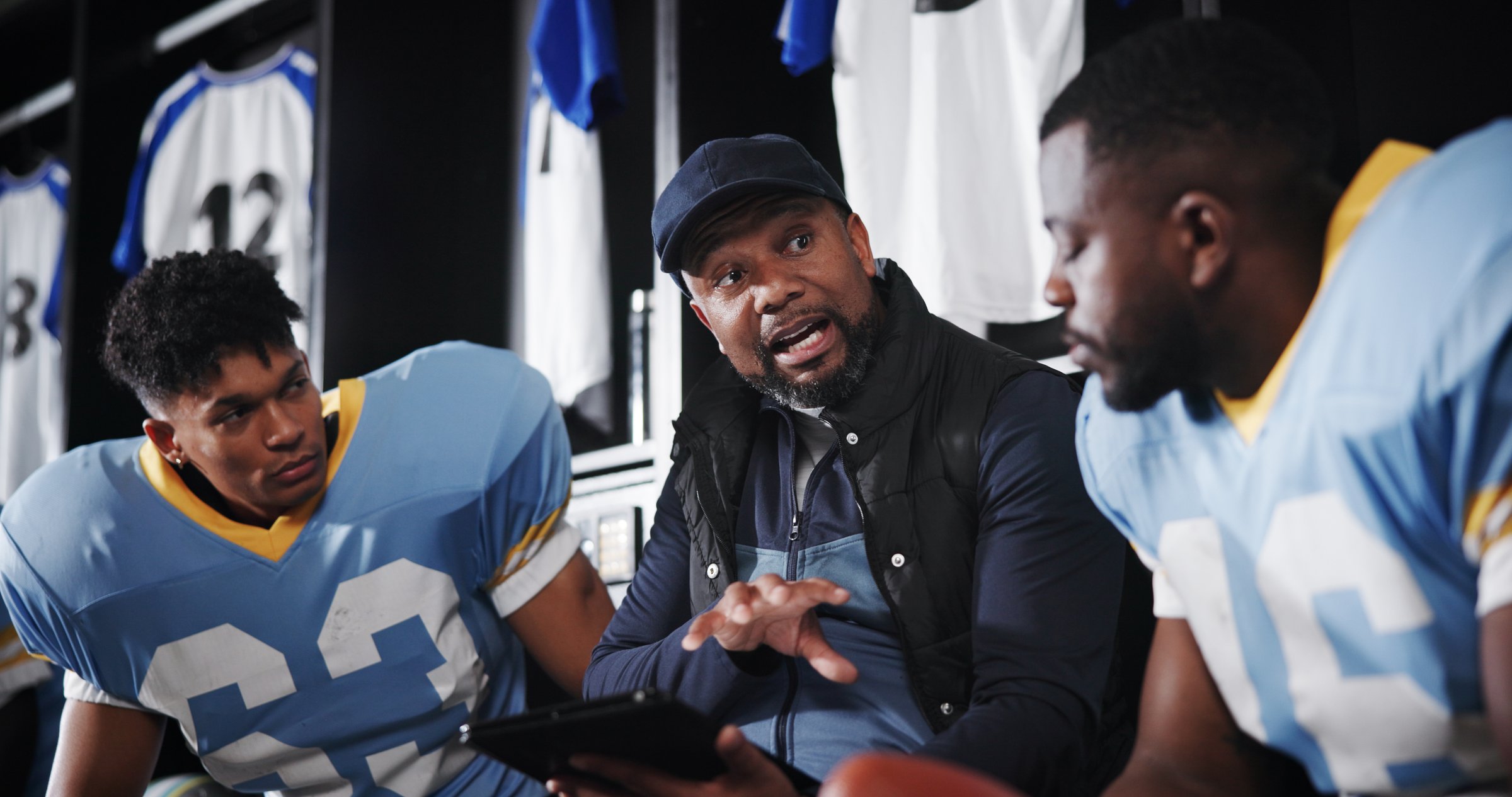 Man, football player and coach with team in locker room, tablet and advice for performance. Mentor, professional athlete and discussion for fitness, sports and planning with strategy at club
