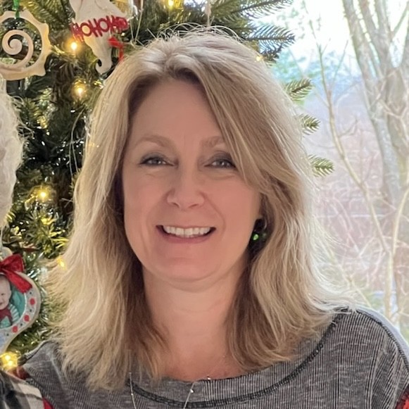 A woman smiling in front of a Christmas tree decorated with ornaments, wearing a gray sweater. Soft outdoor lighting.