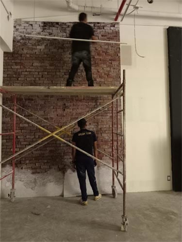 Two workers applying a brick-patterned wallpaper on a large wall using scaffolding in an indoor setting.