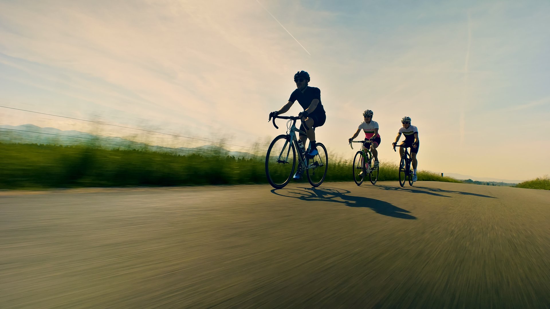Three cyclists pedal down a scenic road, their shadows stretching behind them. The motion blur conveys speed and energy, emphasizing the thrill of road cycling. Aerial view.