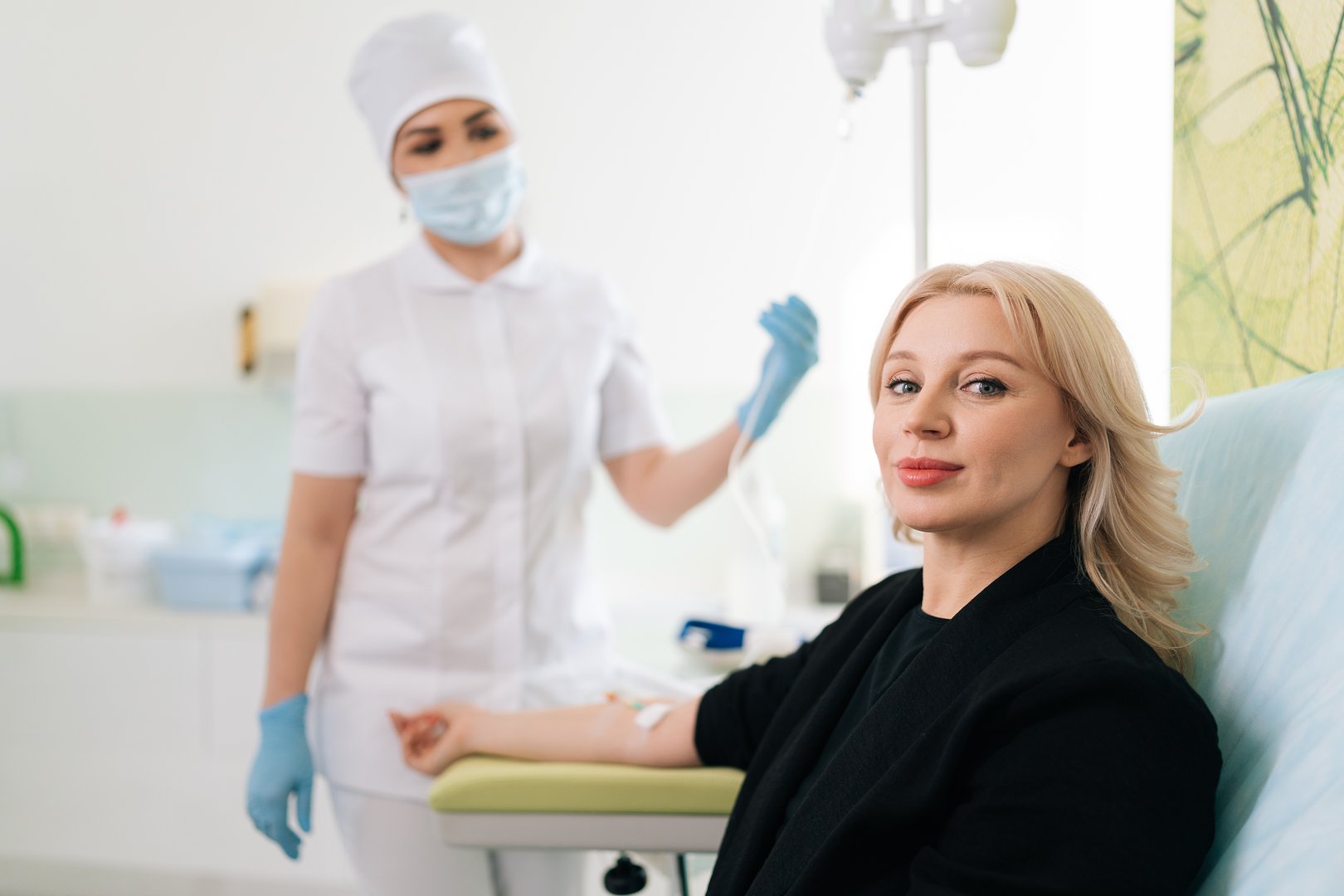 Portrait of blonde woman sitting on medical chair and getting intravenous vitamin therapy looking at camera. Clinic nurse in uniform standing on blurred background. Healthcare, wellness concept.