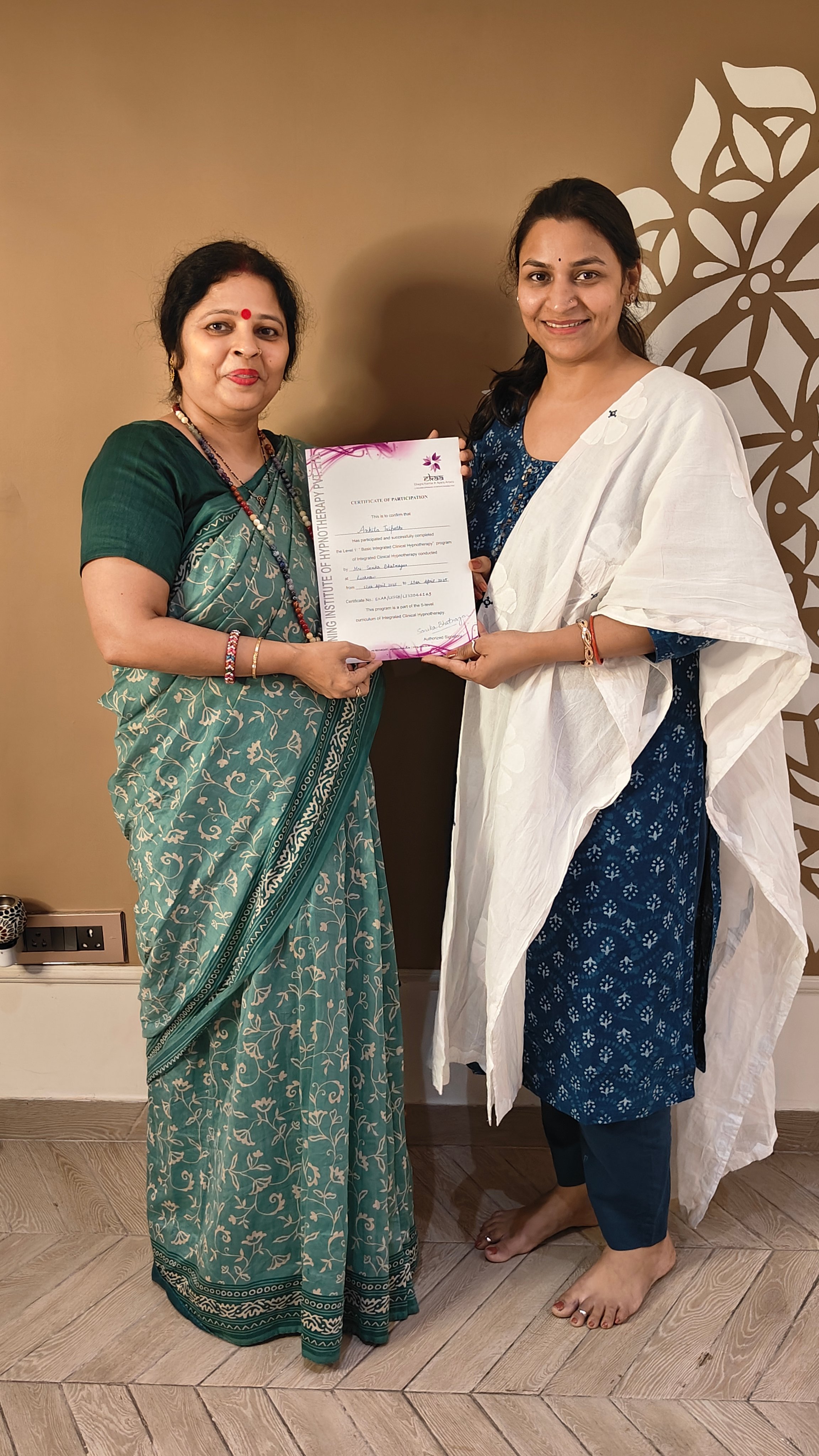 Two women smiling and holding a certificate, standing in front of a wall with floral patterns.