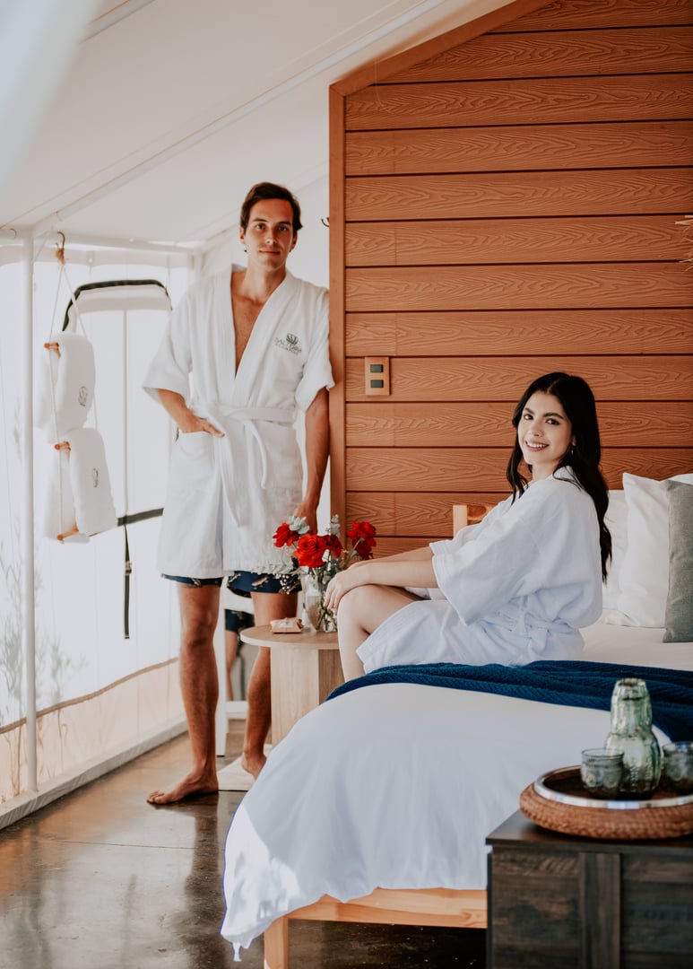 Two people in white robes, one standing and one sitting on a bed in a cozy wooden room with flowers on a table.