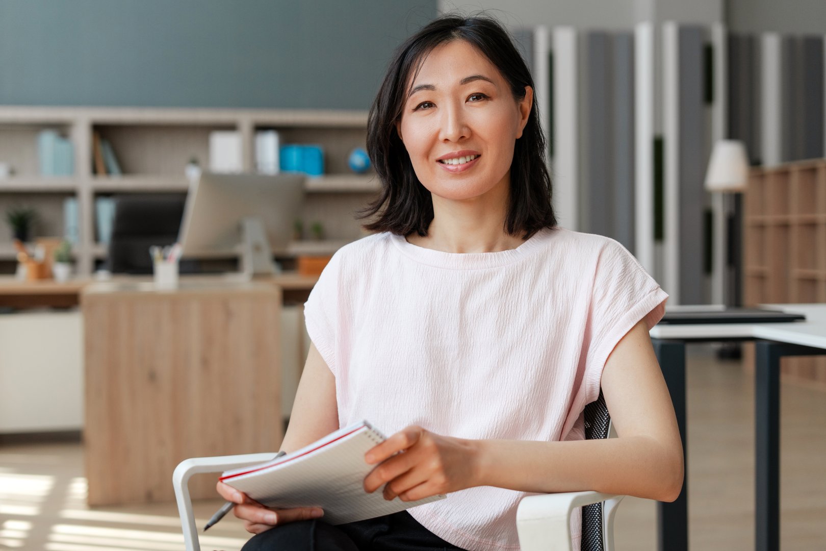 Attractive Asian businesswoman is sitting in her office, holding a notebook and smiling. Successful business concept