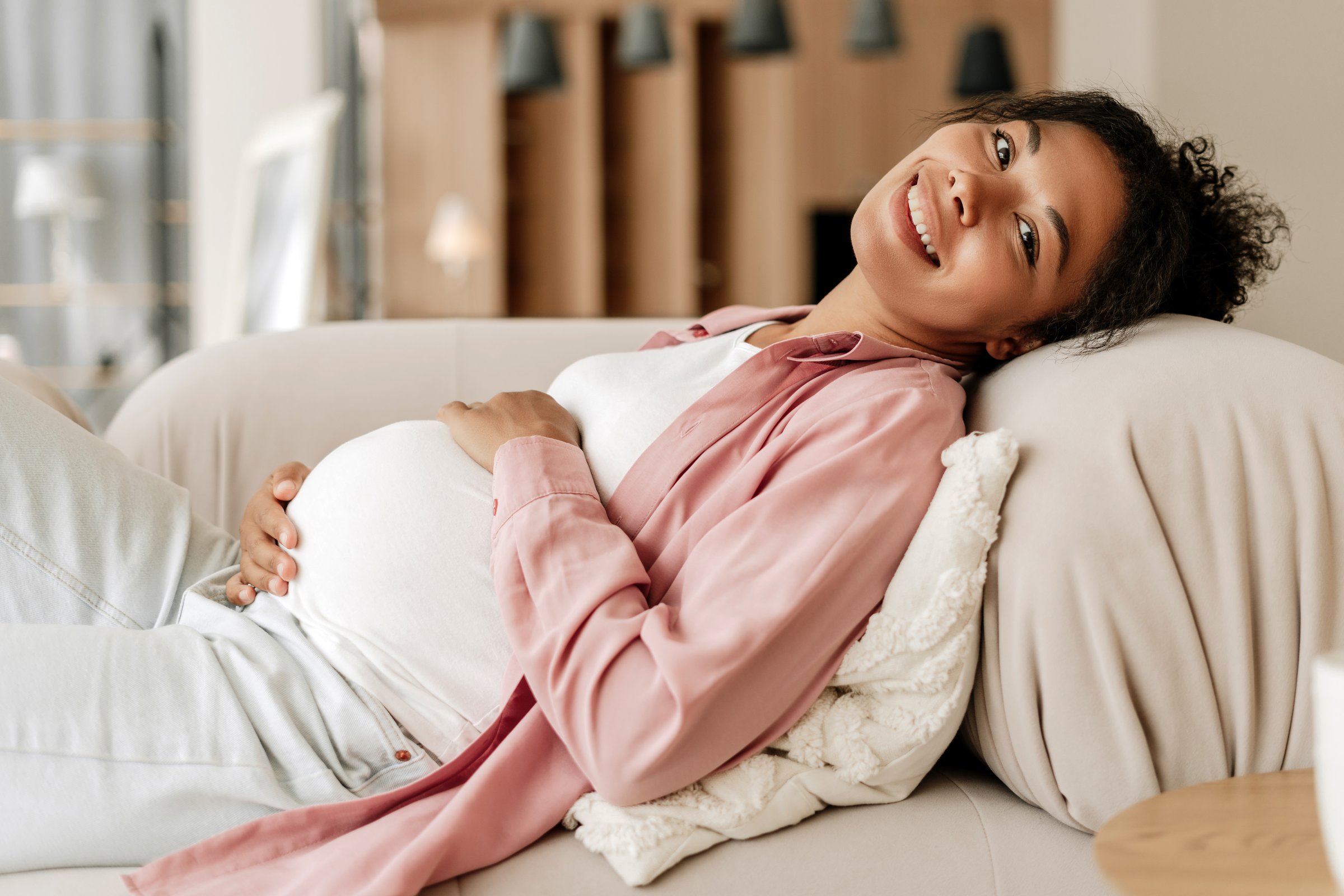 Pregnant woman is lying on a sofa with her hand on her belly, smiling serenely