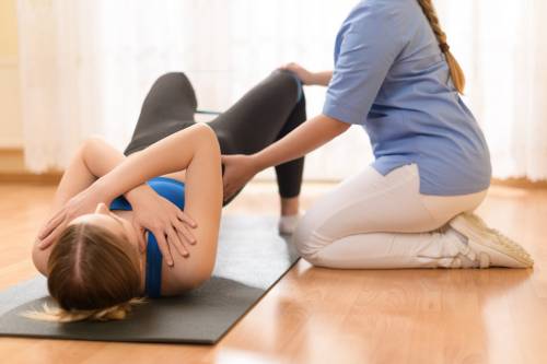 Woman on mat being assisted by a physical therapist in performing a bridge exercise in a rehabilitation setting.