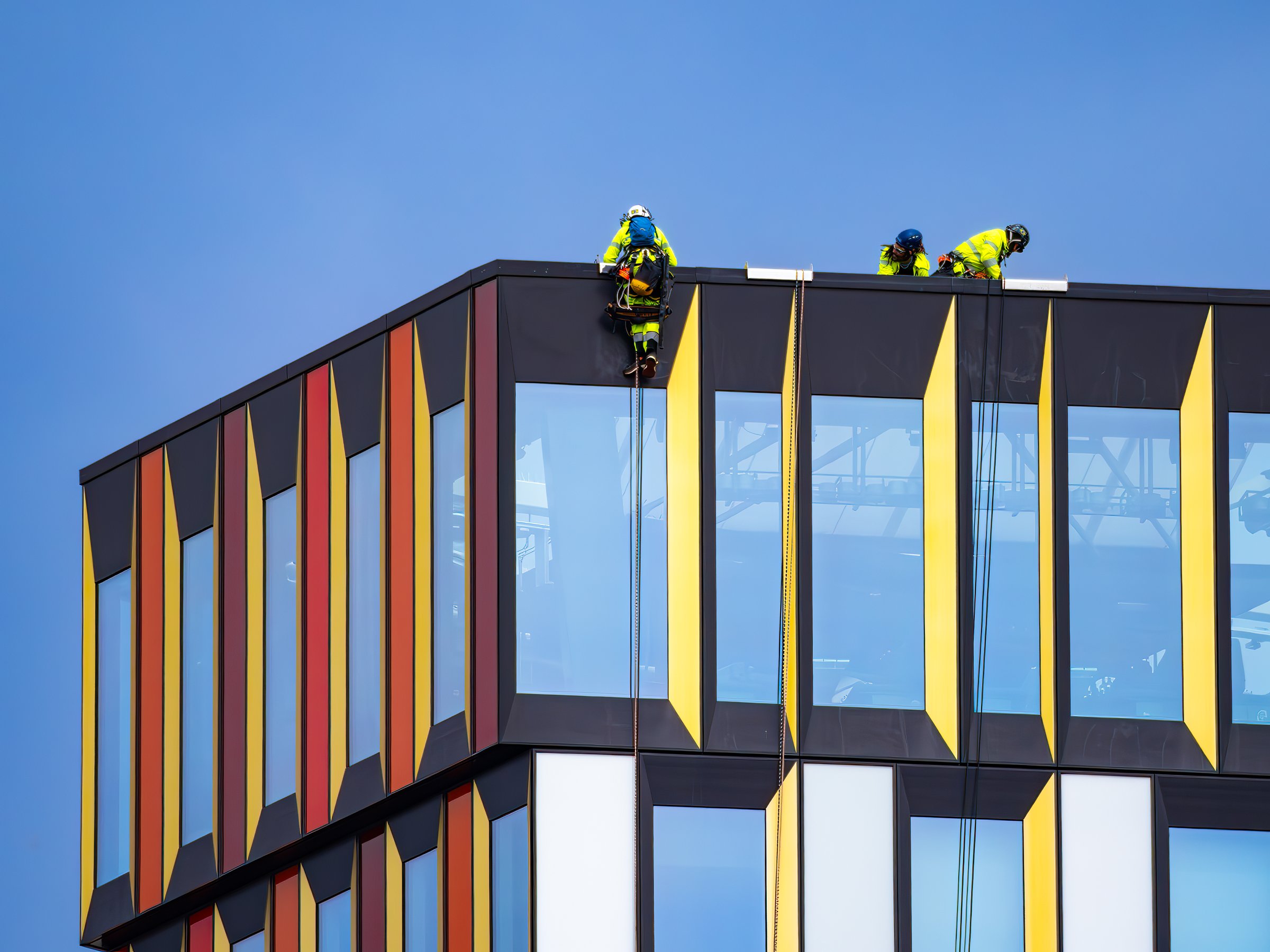 Stockholm, Sweden - June 10, 2025: Workers in safety gear performing maintenance on a high-rise building. They are using ropes to descend from the rooftop, showcasing a modern architectural design.