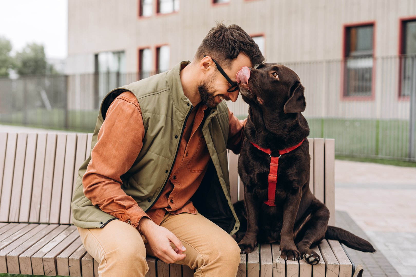 Happy Latin bearded man wearing eyeglasses embracing with beautiful black labrador, sitting together on the street. Pet love concept