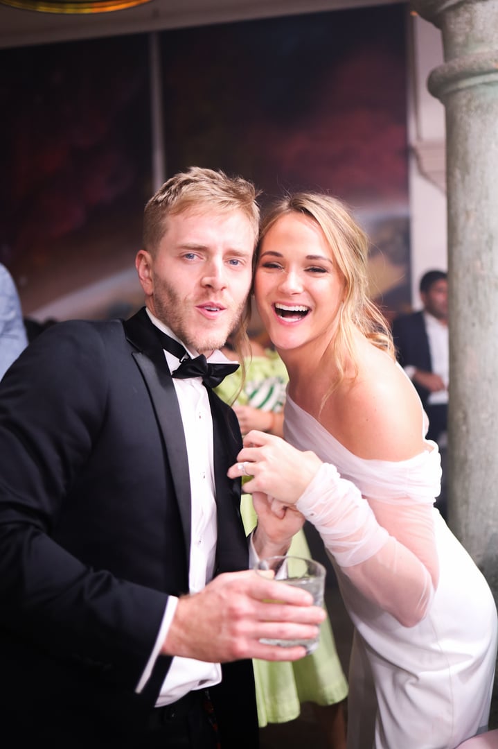 A man in a tuxedo and a woman in a white dress smiling at a party, with him holding a drink, in a festive atmosphere.