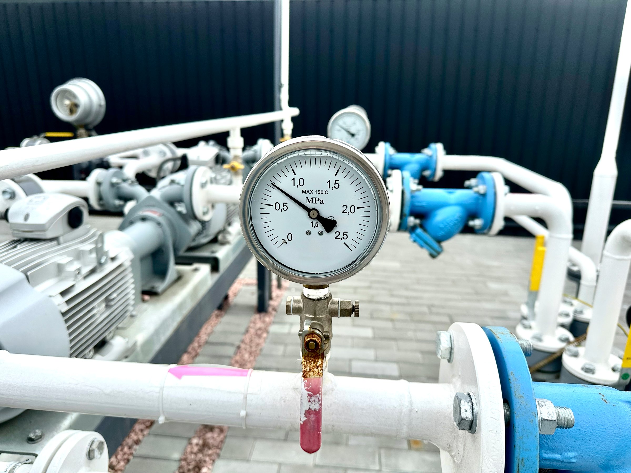 Close-up of industrial pressure gauges connected to white gas pipes at a vehicle fueling station. Equipment used to monitor gas pressure, with visible valves and connections in outdoor setting.
