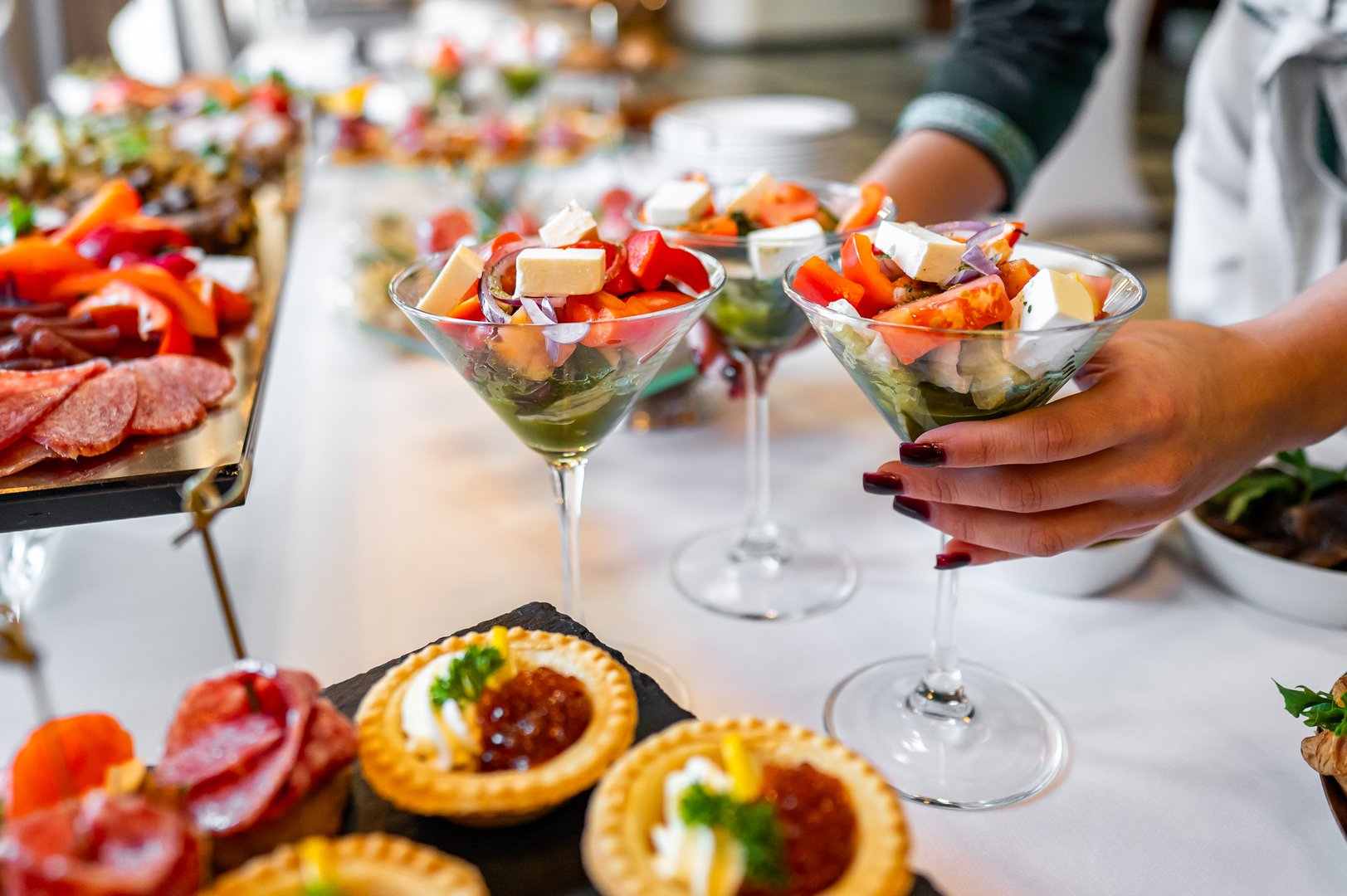 A close-up of a buffet table with an array of gourmet appetizers, including martini glasses filled with fresh salad and tartlets topped with cream and fruit, in a party setting.