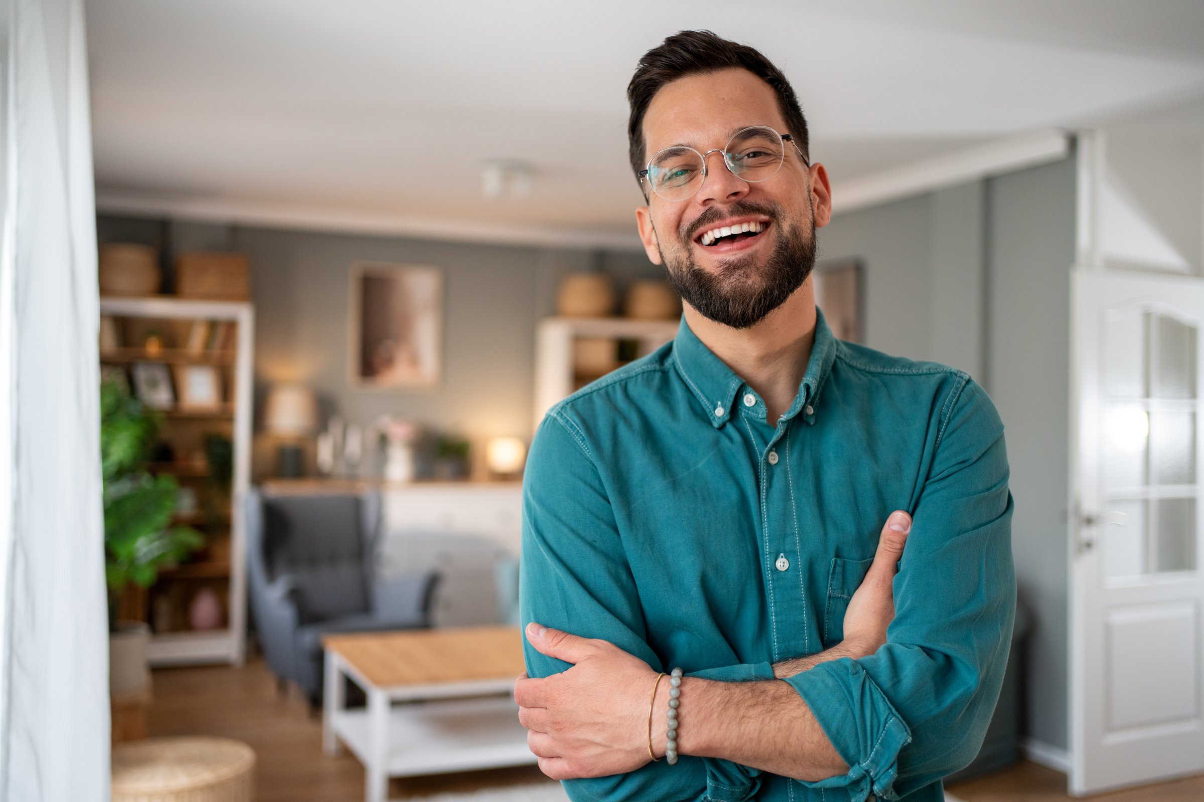 Young man smiling with crossed arms in a modern living room, radiating confidence and happiness. Bright, airy space filled with cozy decor and natural light enhances the relaxed atmosphere