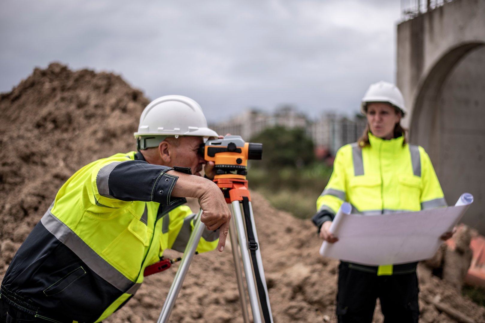Construction workers wearing safety helmets and at a construction site. One worker is using a theodolite (or surveying equipment) on a tripod to take measurements, while the other worker holds a blueprint or set of construction plans.