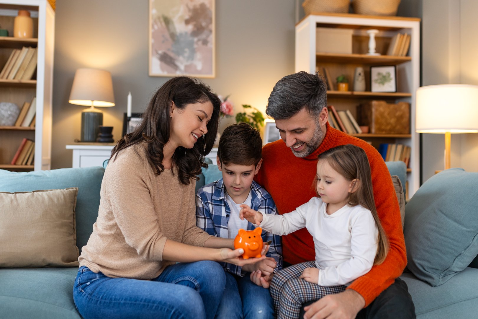 Happy family cheerful mother and father with kids smiling and putting coins into piggy bank while sitting on sofa at home
