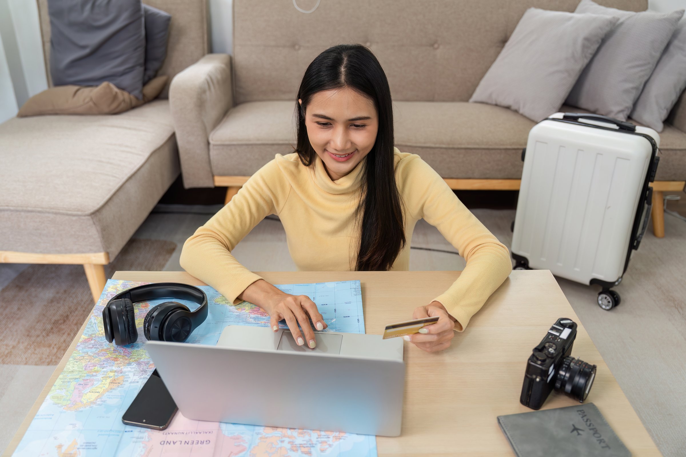 A young woman books accommodation online using her laptop and pays with a credit card, sitting in a modern living room with travel essentials around her.