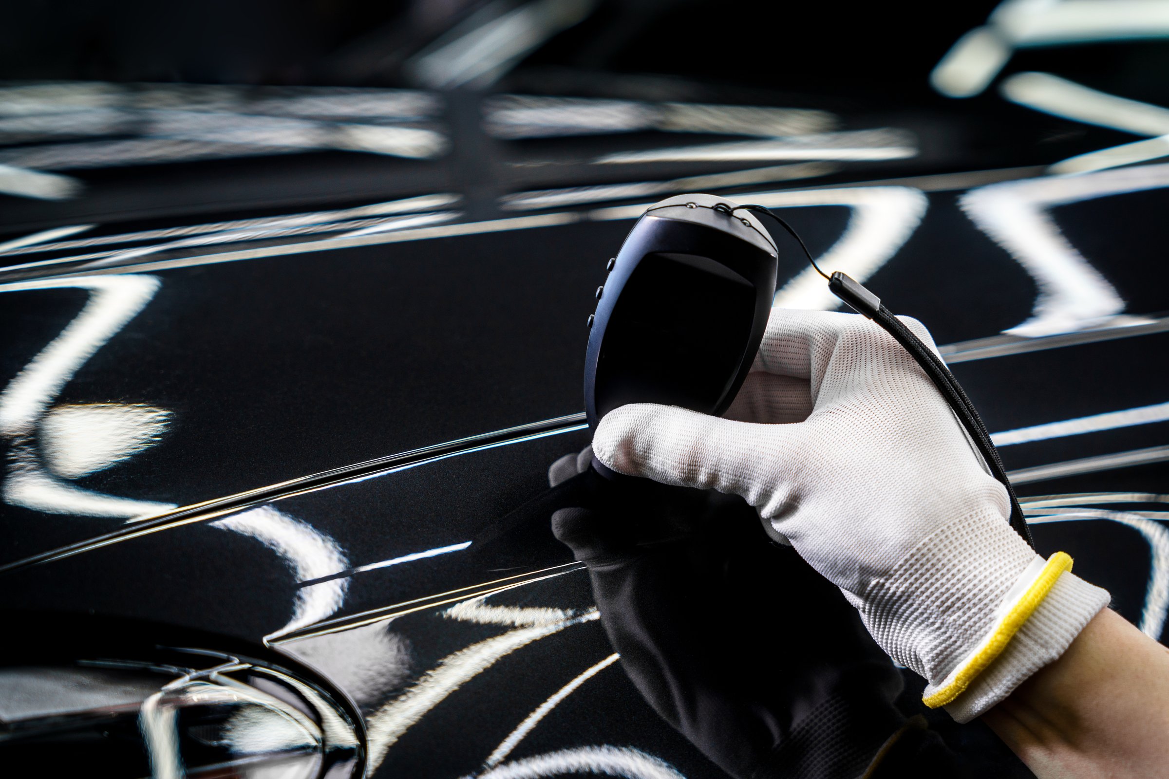 Technician measures car paint thickness with a specialized device on glossy black surface, ensuring quality and uniformity in automotive finishing