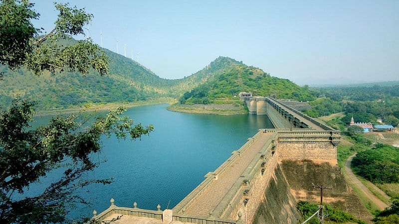 A scenic view of a large dam with blue water, surrounded by green hills and trees under a clear sky.