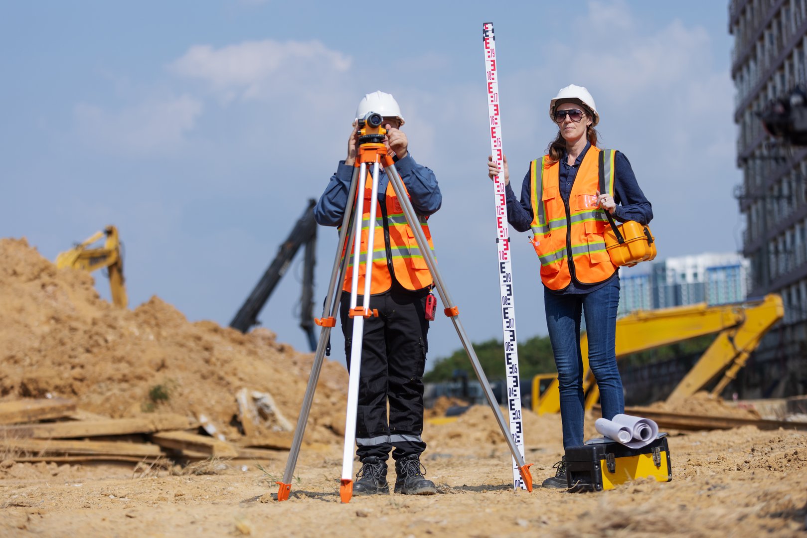 Two construction surveyors in safety gear perform land measurement at a worksite using a total station and leveling rod, surrounded by dirt piles, excavators, and construction equipment.