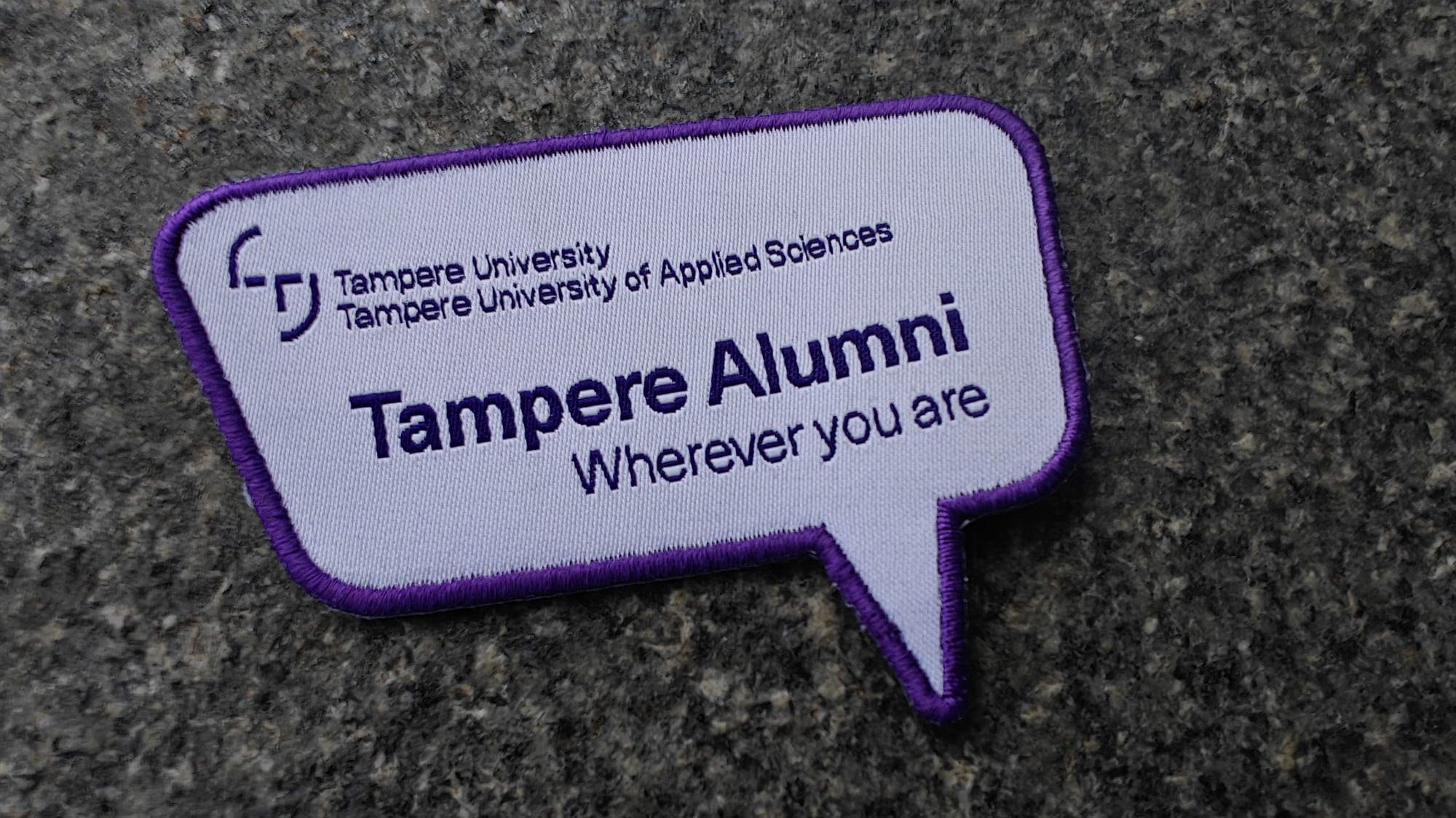A speech bubble patch with Tampere Alumni Wherever you are and logos for Tampere University on a gray surface.