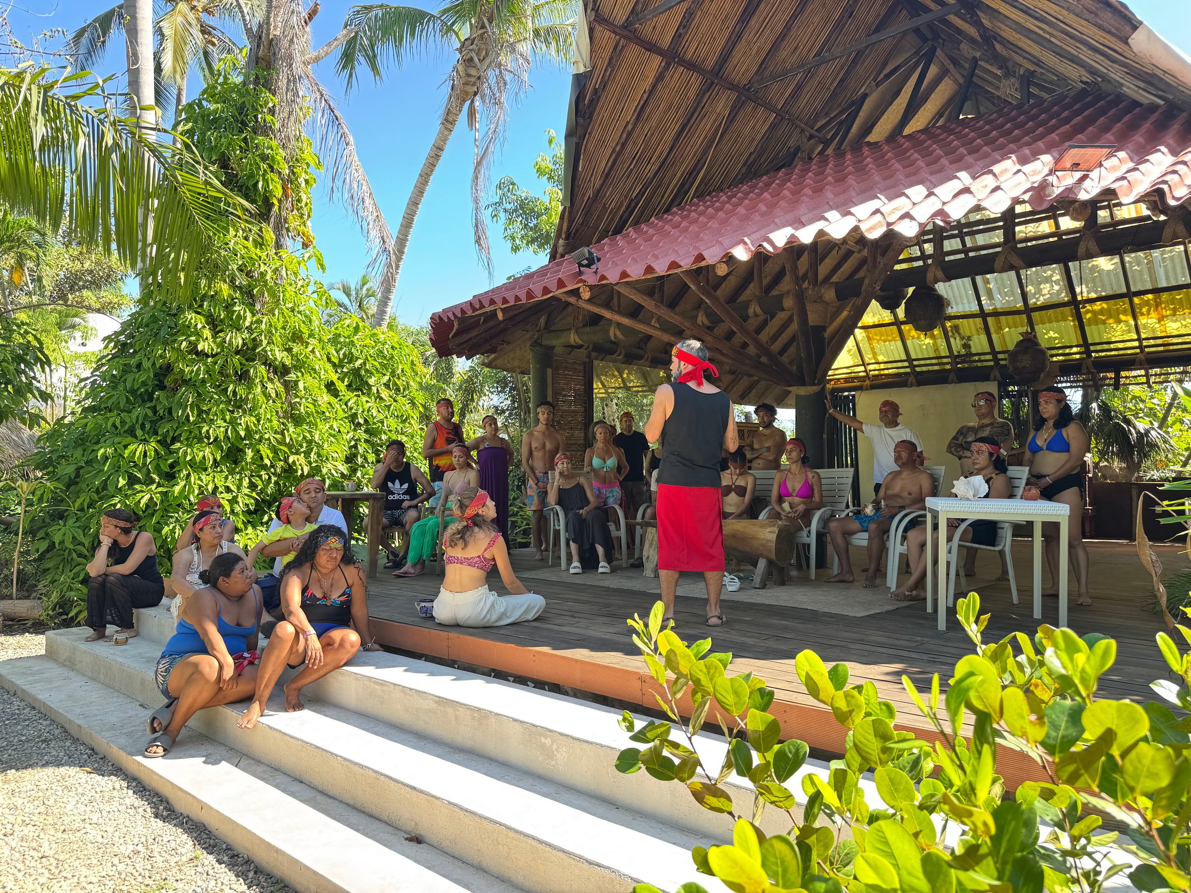 A group of people attentively listen to a speaker under a tropical pavilion with lush greenery and palm trees around.