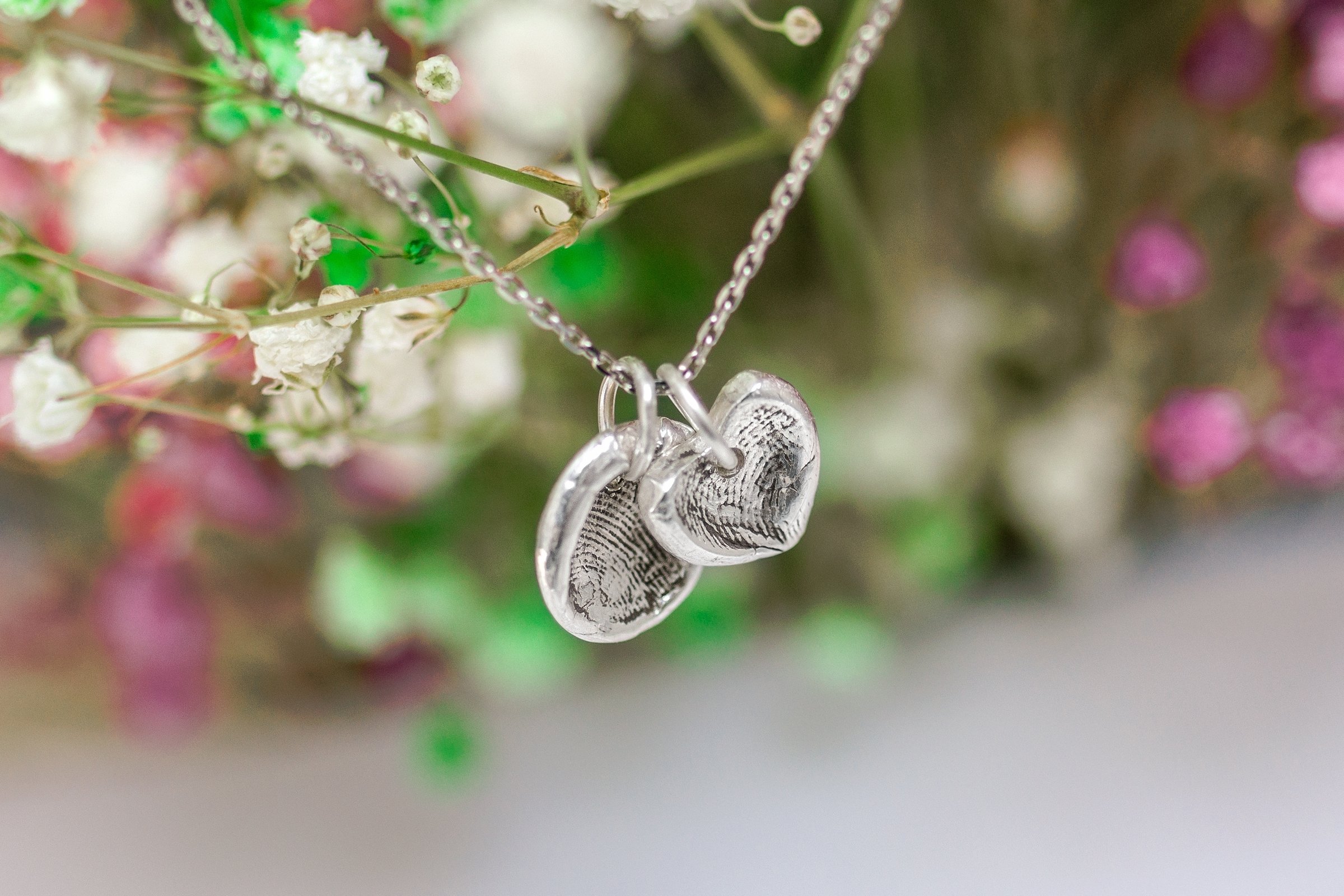 Delicate Silver Necklace with Heart and Round Pendants Featuring Fingerprint Details Amidst Flowers.