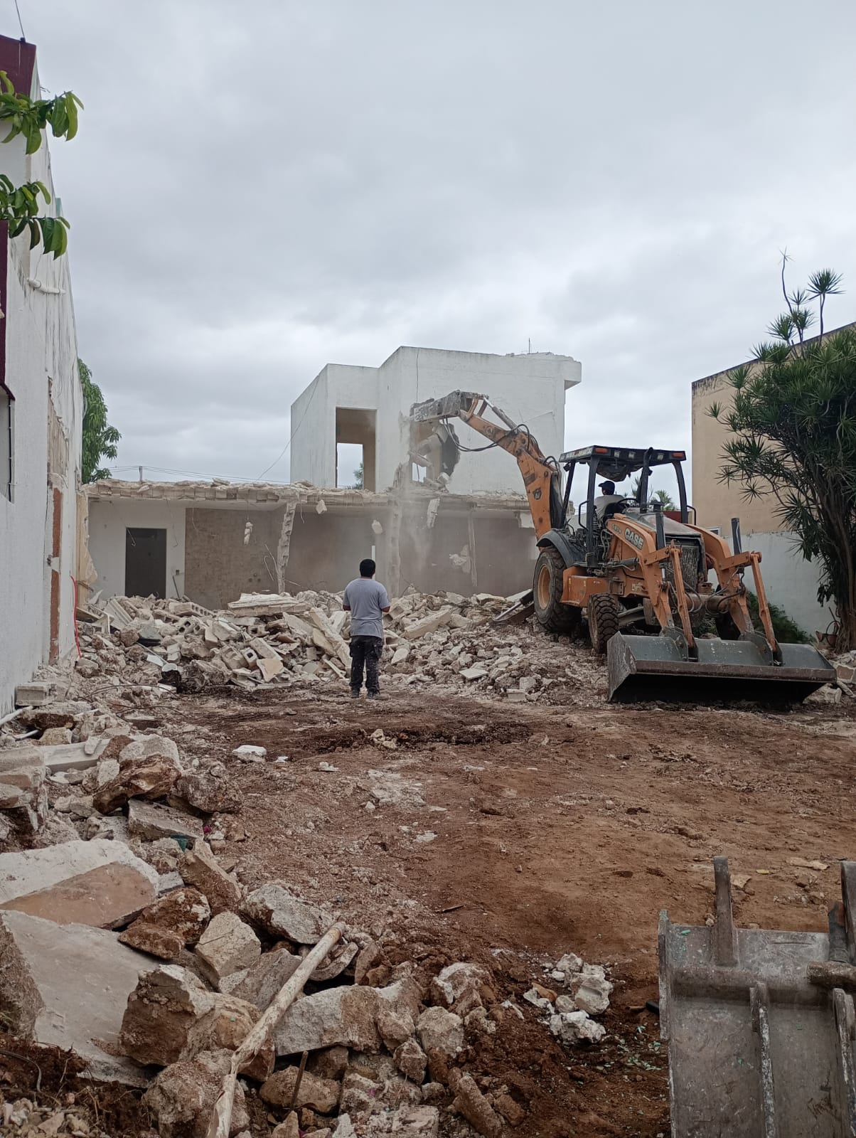 A bulldozer demolishing a building as a person looks on, surrounded by debris, under a cloudy sky.