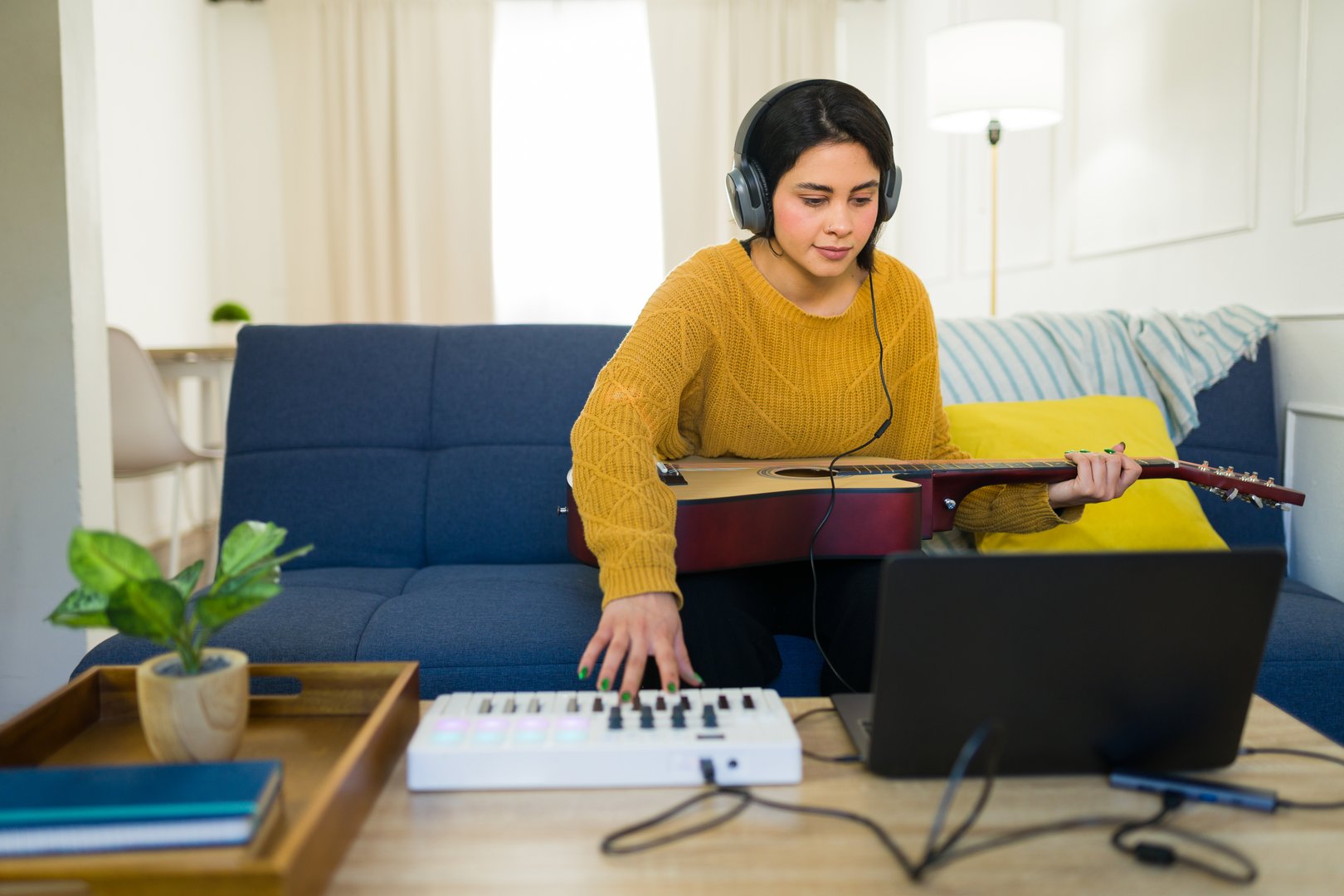 Latina musician creating music, playing guitar and midi keyboard while working on laptop in home studio setting