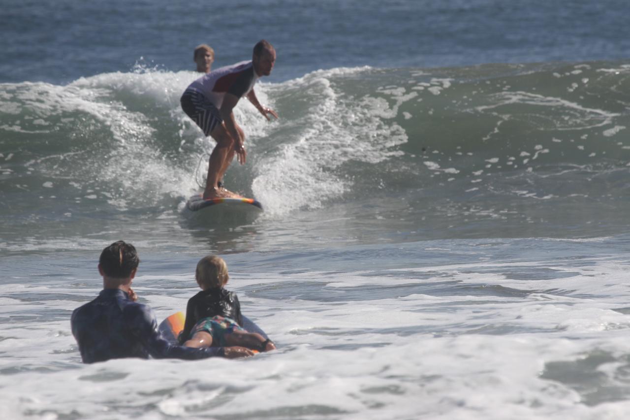 Surfer riding a wave while two people watch from the water, with a clear blue ocean and sky in the background.