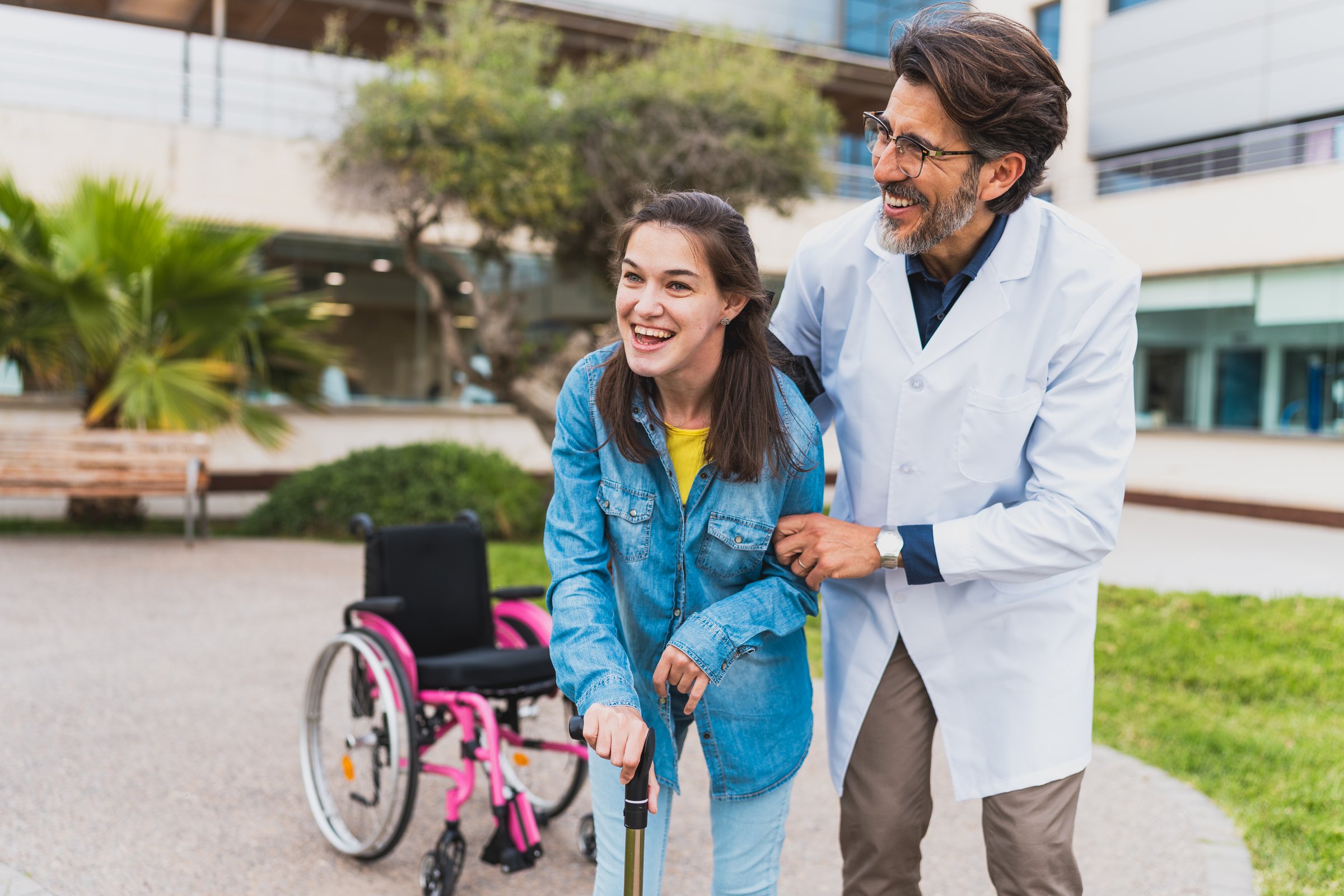 Smiling young woman with cerebral palsy walks with a cane, helped by a doctor. Concept of cerebral palsy.