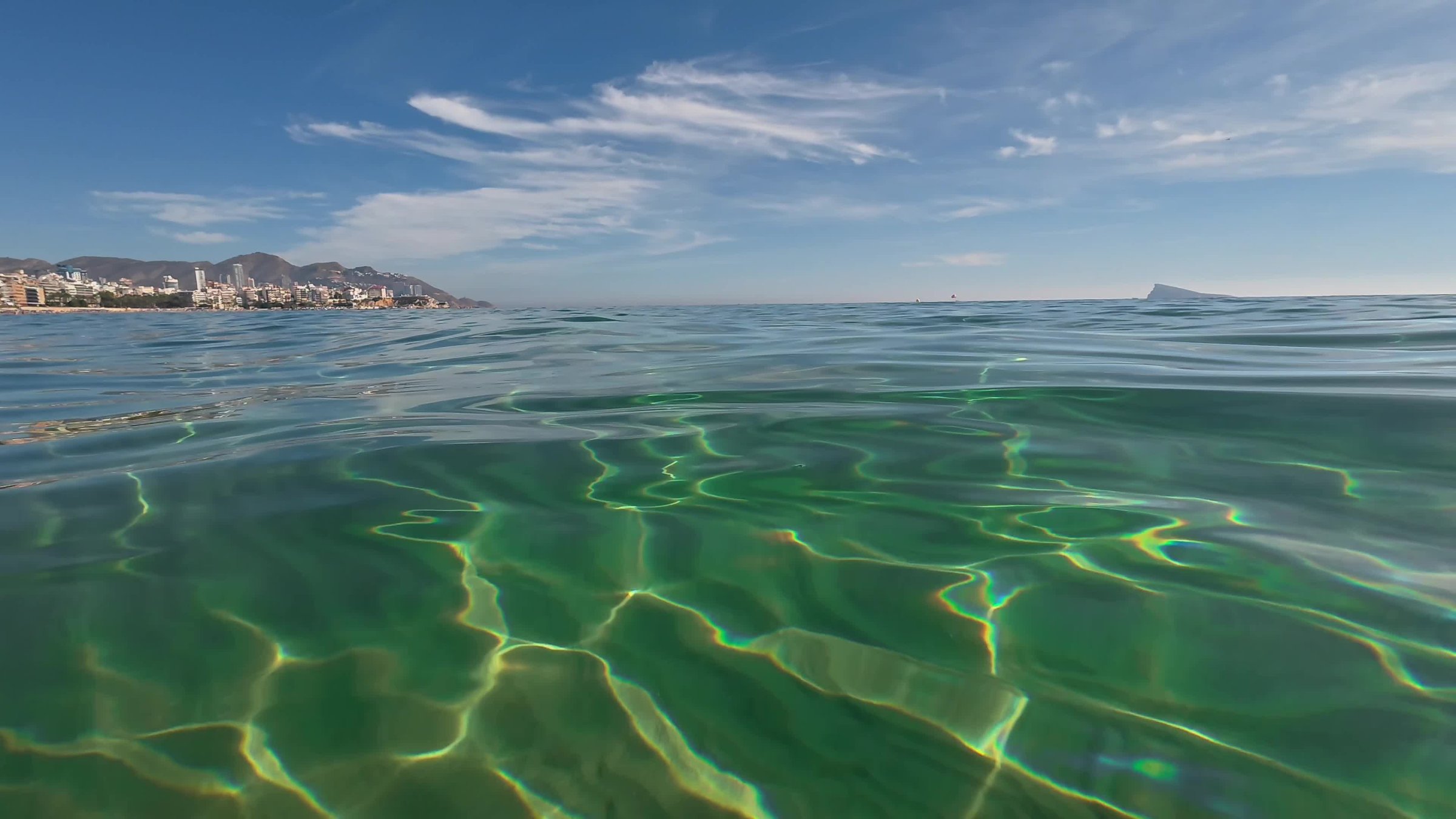 Shallow Emerald Water Rippled Sand Shoreline, Glassy Surface With Sunlight Patterns On Seabed, Distant Coastal