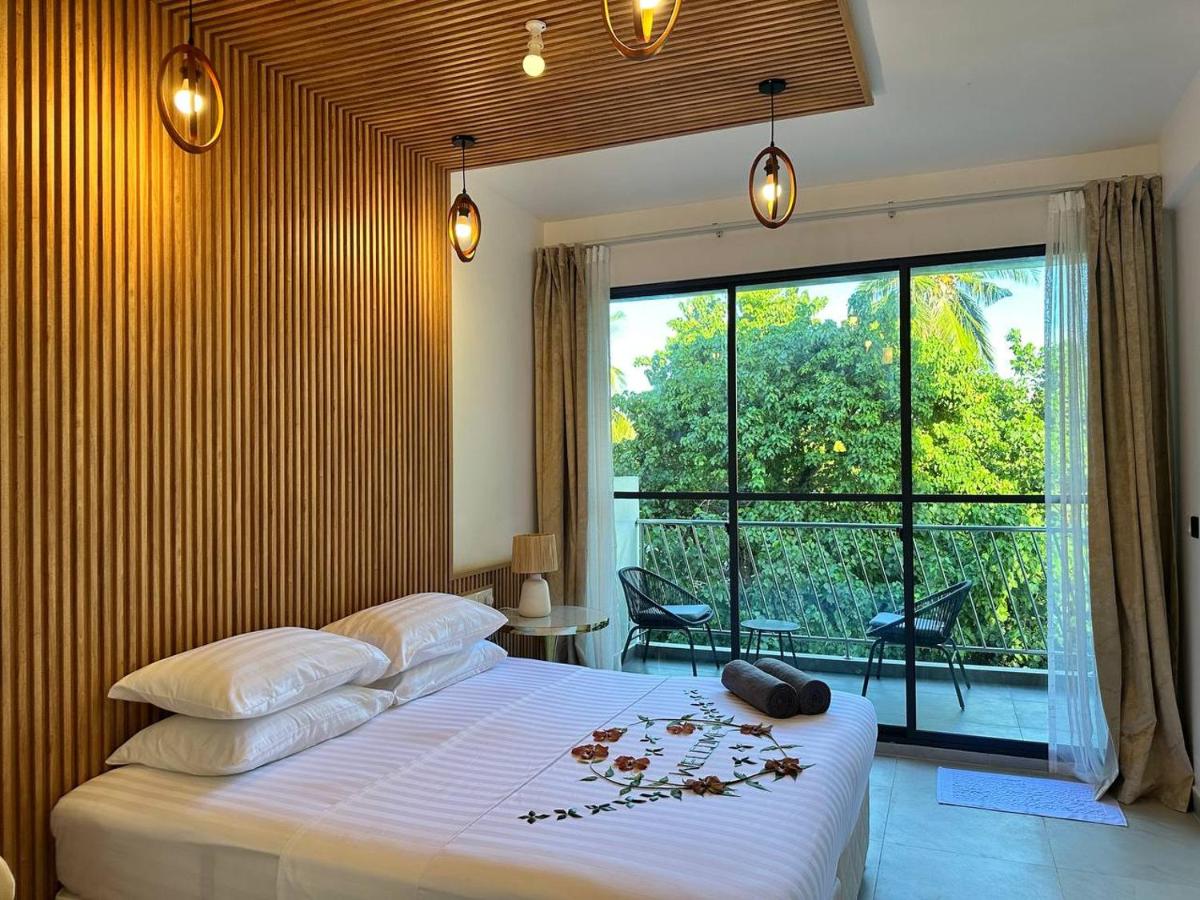 Modern bedroom with wooden accent wall, large window, and balcony view of lush greenery. Bed decorated with flower petals.