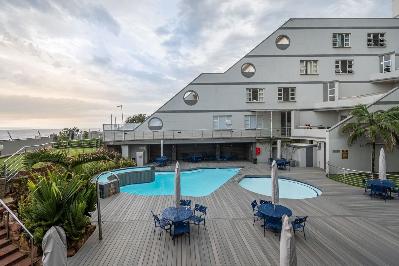 Modern hotel pool area with umbrellas, chairs, and a unique building design under a cloudy sky.