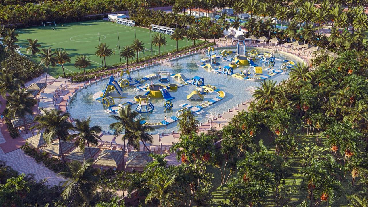 Aerial view of a tropical water park with inflatable obstacles and palm trees, adjacent to a soccer field.