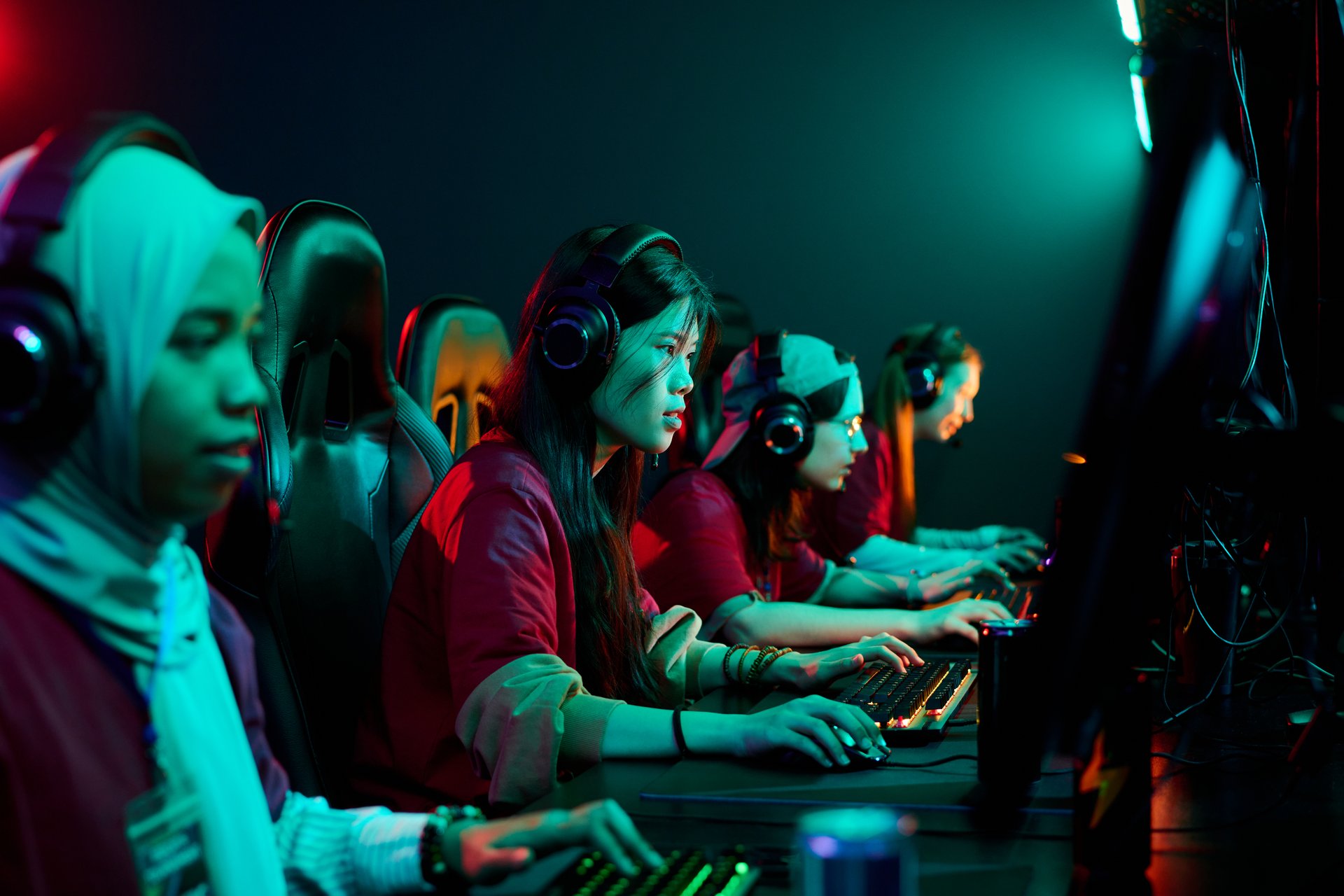 Diverse group of teenage girls wearing headsets competing in esports tournament, sitting in row at gaming computers, focused on screens, illuminated by colorful lights, team gaming together