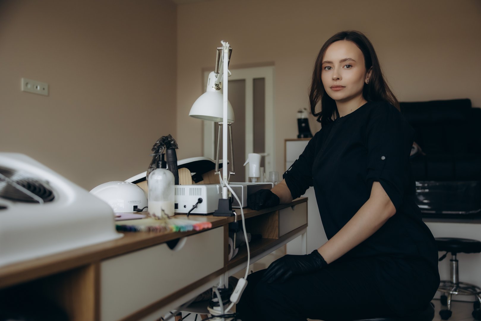 Manicurist wearing black uniform and gloves, posing confidently at personal workspace with nail tools