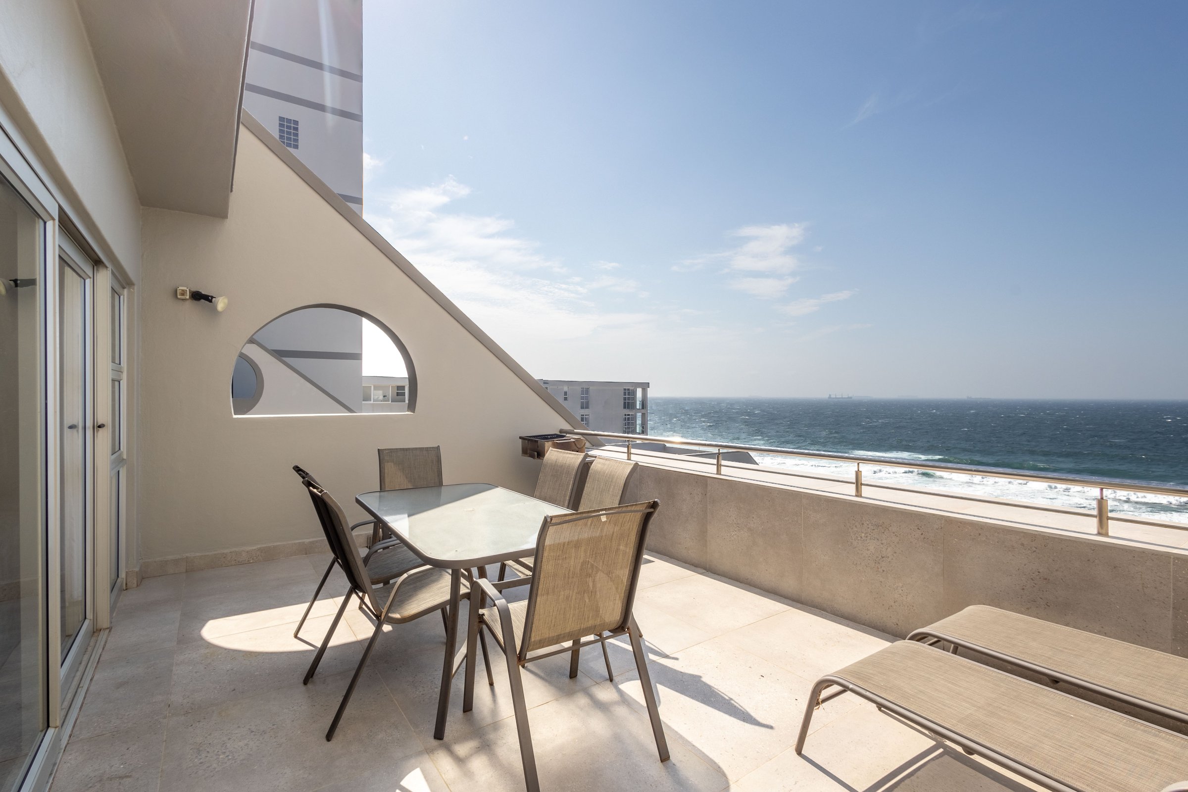 Beachfront balcony with a glass table, chairs, and loungers overlooking the ocean under a clear blue sky.