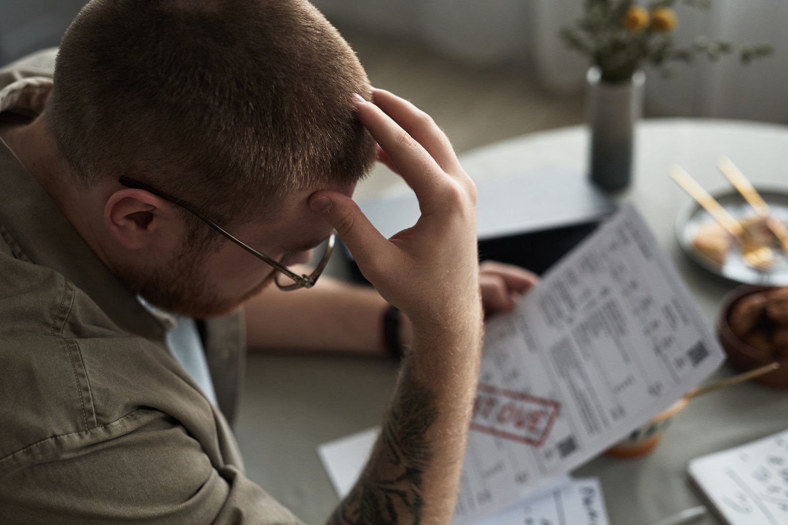 Caucasian young adult man sitting at table holding overdue bills, looking stressed and frustrated, hand on forehead, financial documents and chopsticks visible on surface