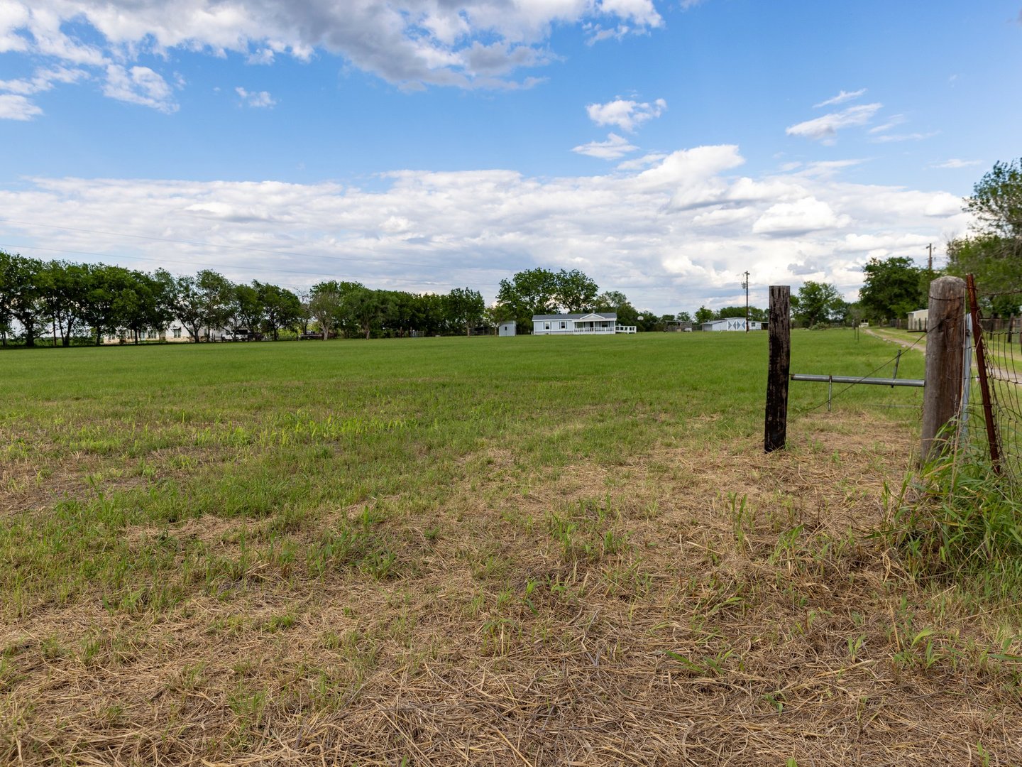 Open grassy field with a wooden fence in the foreground and a few small houses in the distance under a partly cloudy sky.