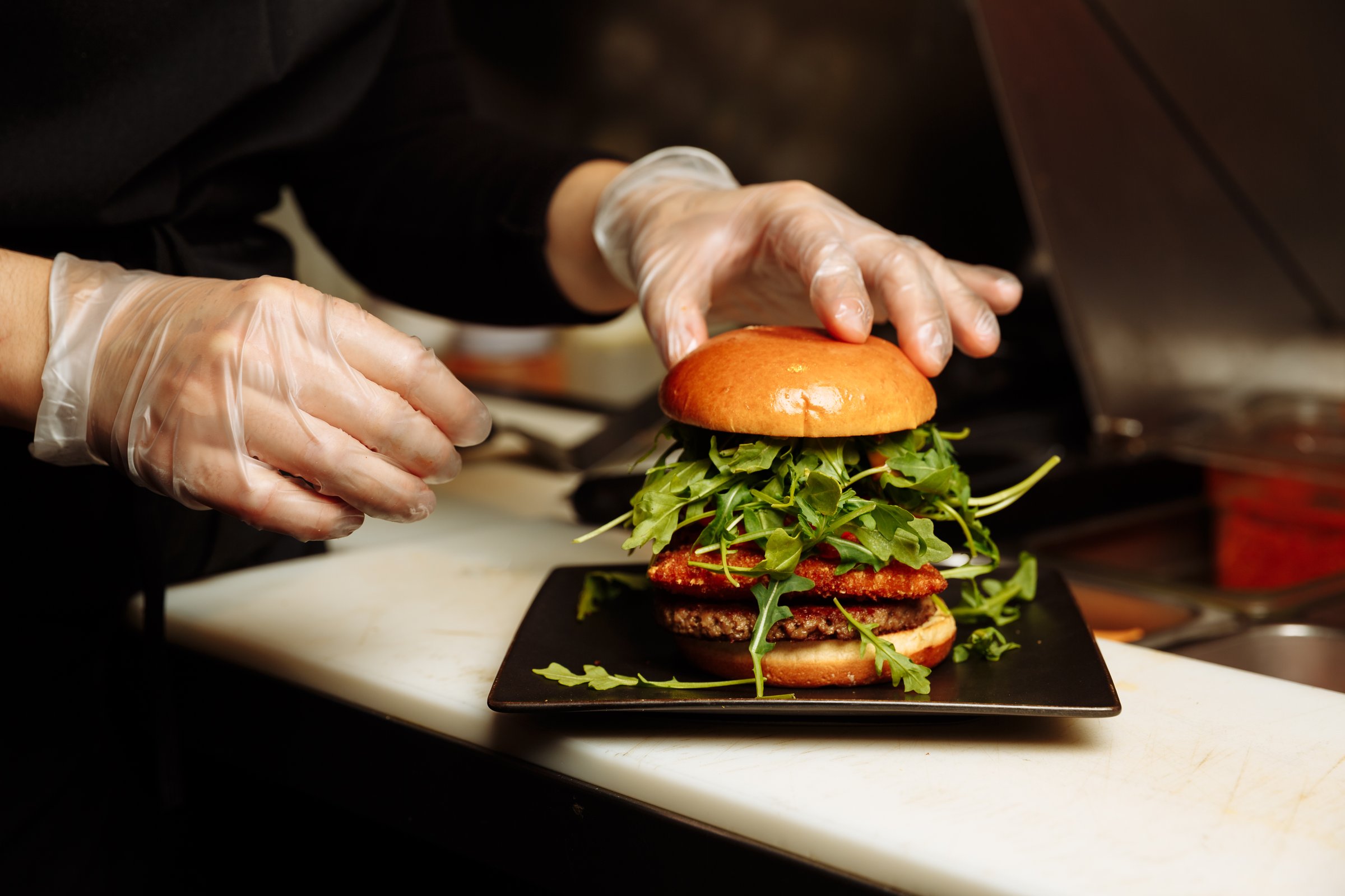 Chef hands in gloves assembling vegan burger with plant based patty and fresh arugula in restaurant kitchen