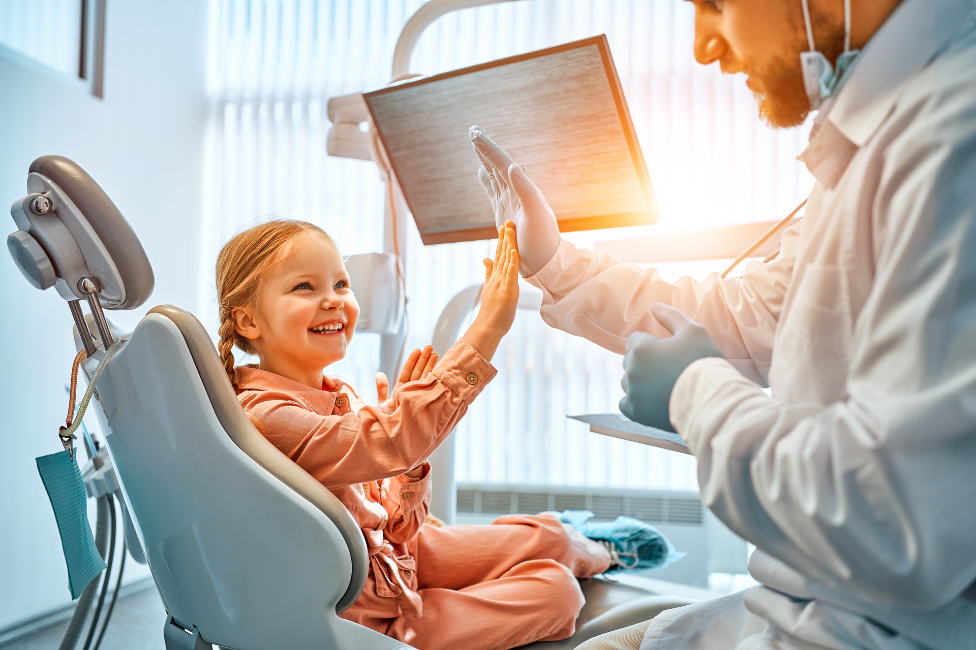 A little girl is sitting in a dentist's chair, giving a high five to the doctor and laughing