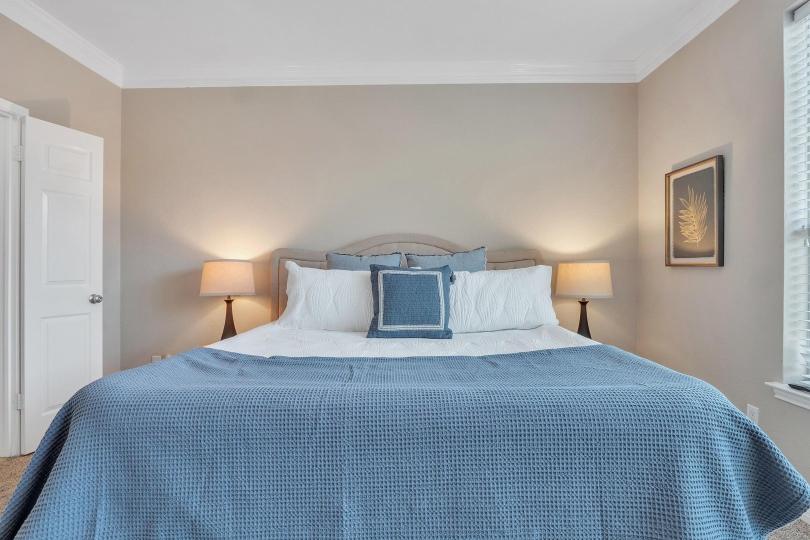 A neatly made bed with a blue blanket and several pillows, flanked by two lamps, in a softly lit bedroom.